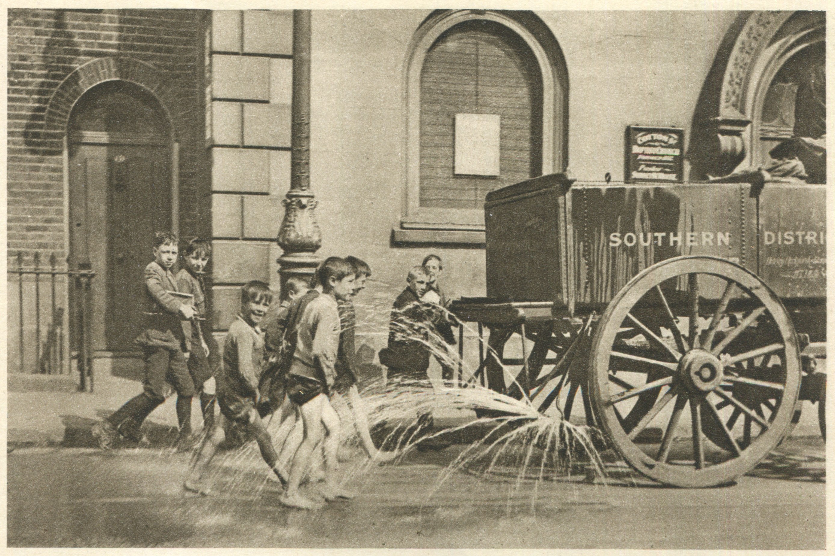 Schwarz-weißes Bild von Kindern, die mit einem Feuerhydranten vor einem Gebäude mit Fenstern, einer Tür, einem Mast und einem Zaun spielen, im Hintergrund ein Wagen.