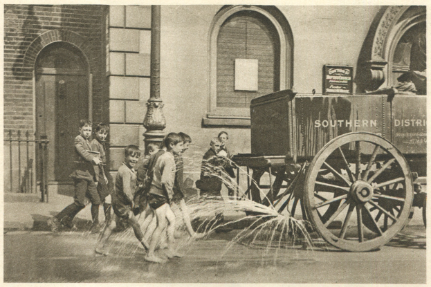 Schwarz-weißes Bild von Kindern, die mit einem Feuerhydranten vor einem Gebäude mit Fenstern, einer Tür, einem Mast und einem Zaun spielen, im Hintergrund ein Wagen.