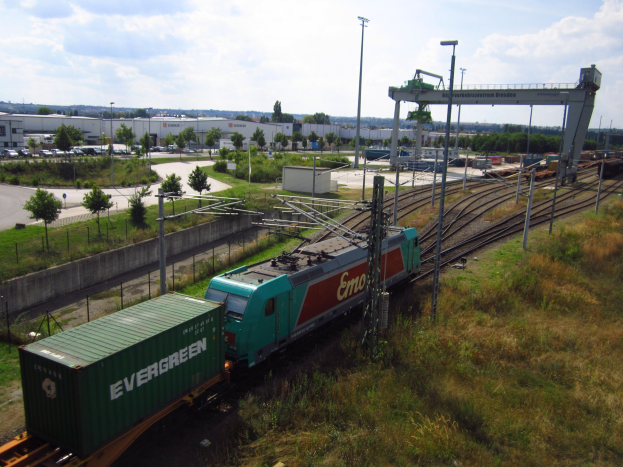 Güterzug fährt auf Schienen neben einem grünen Feld mit Strommasten, Kabeln, Bäumen, Gebäuden, Fahrzeugen und einem bewölkten Himmel.
