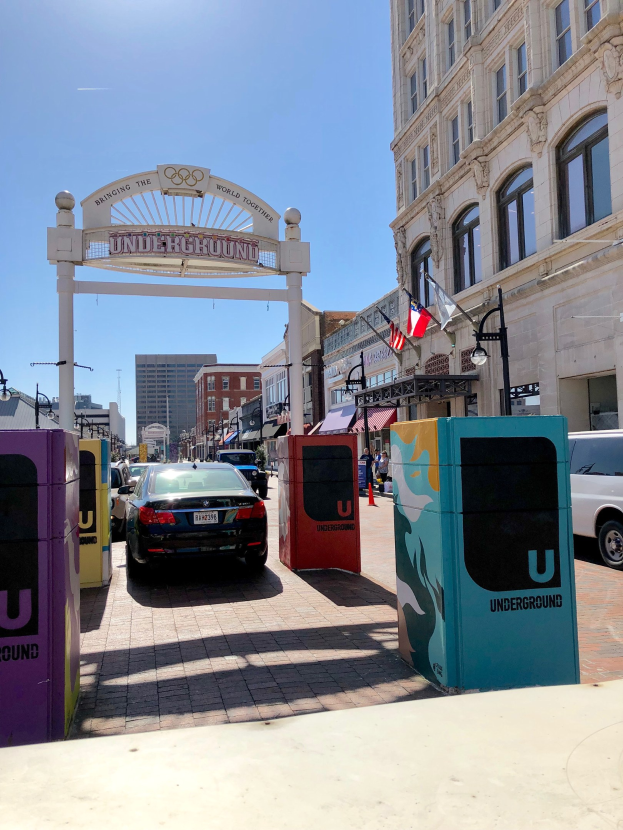 Stadtstraße mit parkenden Autos, einem Torbogen, Gebäuden, Laternen, Fahnen, einem Müllbehälter und Himmel im Hintergrund.