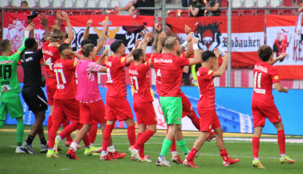 Gruppe von Fußballspielern, die ein Tor feiern, auf einem Feld mit Flaschen haltenden oder klatschenden Spielern, mit Bannern, einem Zaun, Metallstangen, Stühlen und einer Decke mit Deckenleuchten im Hintergrund.