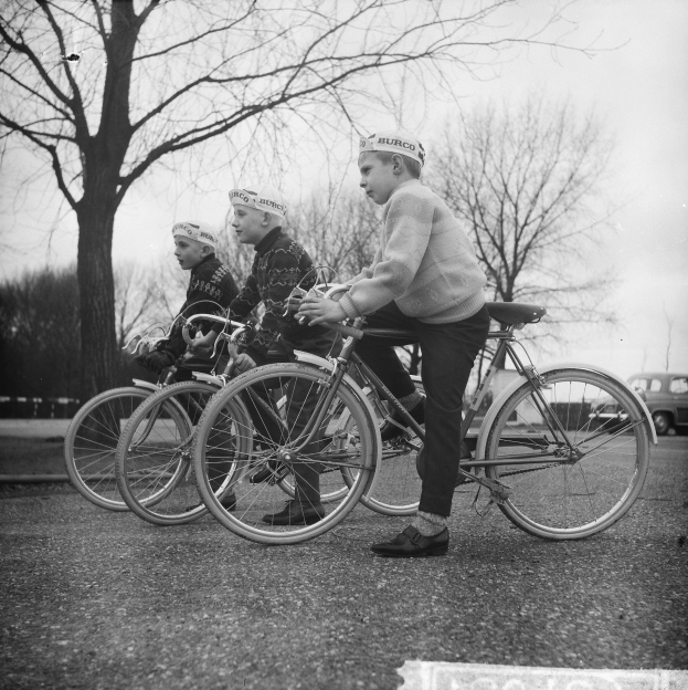 Drei Jungen auf Fahrrädern die Straße entlang mit einem Auto im Hintergrund, in Schwarz-Weiß.