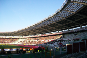 Ein großer Stadion mit vielen Zuschauern bei einem Fuballspiel, mit Menschen, die stehen und sitzen, einem Zaun, einem Schild, Pfosten, einem Dach mit Deckenleuchten und einem bewölktem Himmel.