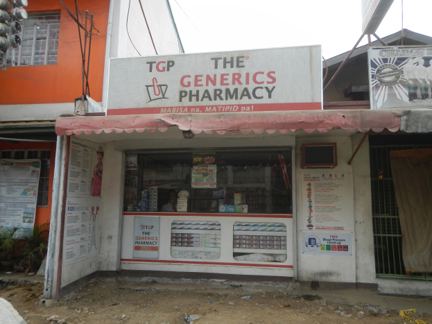 Außenansicht des 'The Generics Pharmacy'-Ladens in einer städtischen Gegend mit umgebenen Gebäuden, Infrastruktur und Fahrzeugen unter einem klaren blauen Himmel.