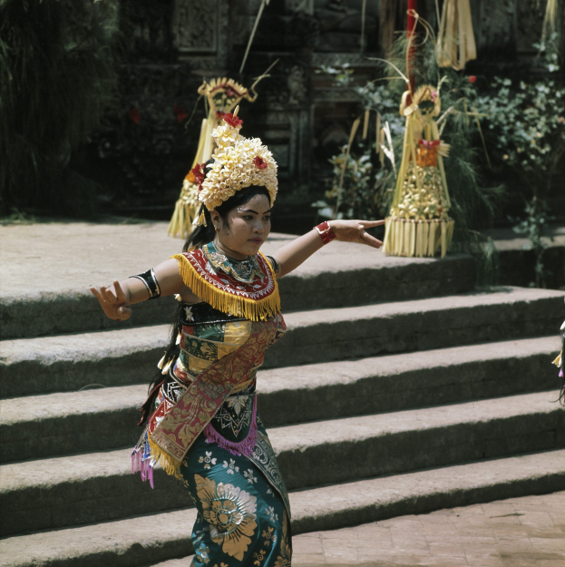 Eine Frau in einem bunten Kleid tanzt auf den Stufen eines Tempels umgeben von Pflanzen und anderen Gegenständen.