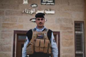 Ein Polizeibeamter in Uniform und Mütze steht vor einem Gebäude mit Türen, Fenstern und Text an der Wand.