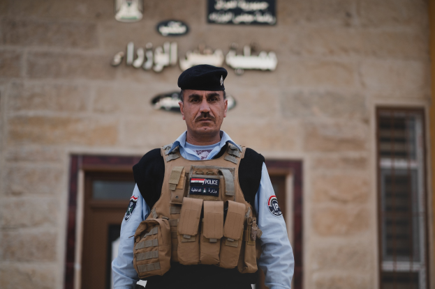 Ein Polizeibeamter in Uniform und Mütze steht vor einem Gebäude mit Türen, Fenstern und Text an der Wand.