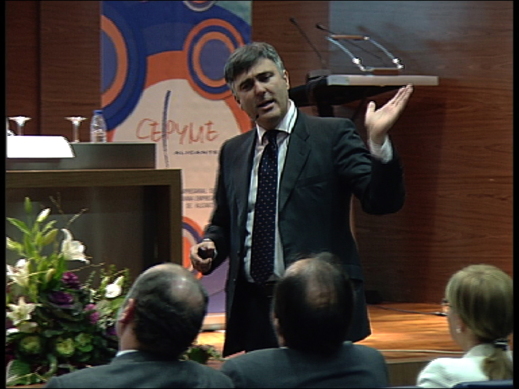 Ein Mann in einem schwarzen Blazer steht an einem Podium mit Mikrofonen, spricht in eines hinein, mit einem Blumentopf oder einem Blumenstrauß daneben und einem Tisch mit einem Buch, einer Wasserflasche und anderen Gegenständen in der Nähe; dahinter eine braune Wand und eine farbenfrohe Tafel mit Text, mit drei Personen, die im Vordergrund auf Stühlen sitzen.