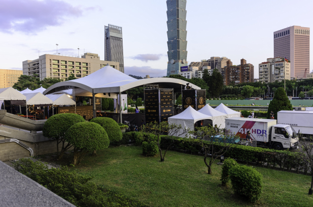 Eine Gruppe von Zelten in einem Park umgeben von Grünanlagen, mit Menschen, Fahrzeugen, Gebäuden und einem klaren Himmel im Hintergrund, möglicherweise aufgenommen auf dem Hong Kong International Film Festival.