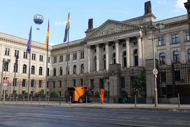 Großes modernes Gebäude mit zahlreichen Fenstern, als Bundestag in Berlin identifiziert, umgeben von Straßeninfrastruktur, Fahrzeugen, Fußgängern und Bäumen unter einem bewölkten Himmel.