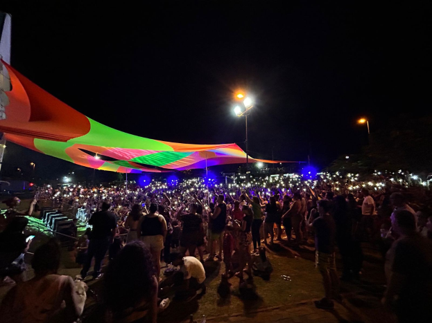 Eine große Menschenmenge steht vor einem Zelt bei Nacht, beleuchtet von bunten Lichtern und Pfählen, mit einem Gebäude links und dem Nachthimmel im Hintergrund, das das 2018er Kunstfest zeigt.