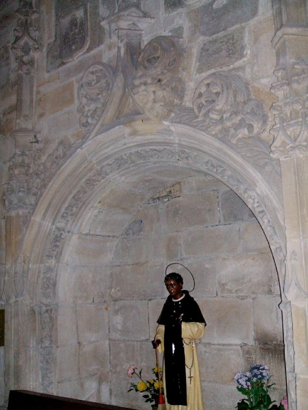 Ein Priester in schwarzer und cremefarbener Robe steht in einem Kirchenbogen und hält einen Stab, mit Topfpflanzen mit Blüten neben ihm und kunstvoll verzierten Wänden dahinter.