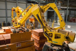 Großes Industriegelände mit gestapelten Kisten, Kränen an der Decke, Zaun, Pfosten und einer fernen Wand in einer automatisierten Fertigungsumgebung.