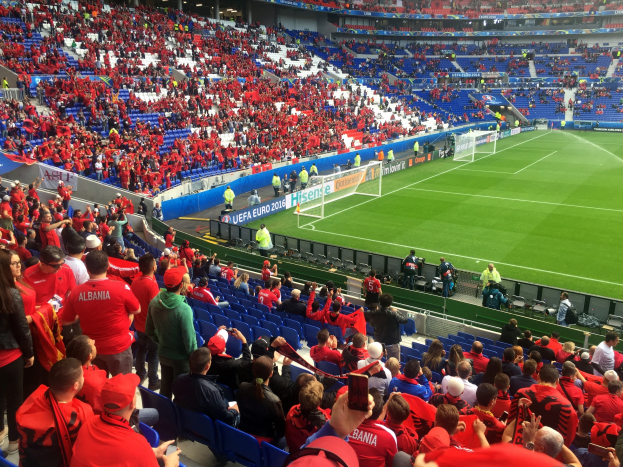 Eine große Menschenmenge in einem Stadion schaut einem Fußballspiel zu, mit Menschen auf Sitzen und Stehplätzen, Schildern, einem Torpfosten und Deckenlichtern.