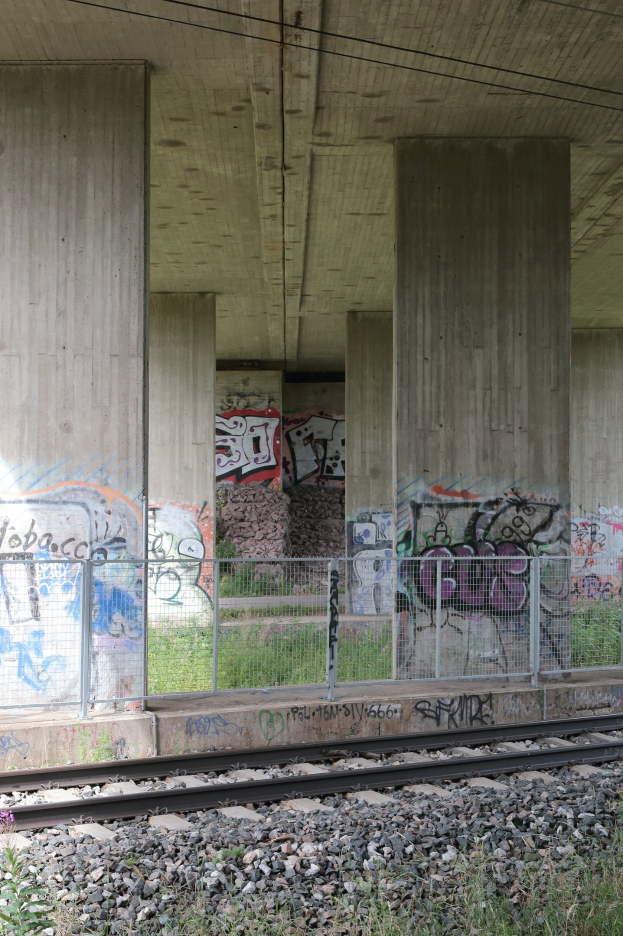 Eisenbahnschiene mit Graffiti an einem Zaun, umgeben von Steinen, Gras, Säulen, Drähten und einem Dach.