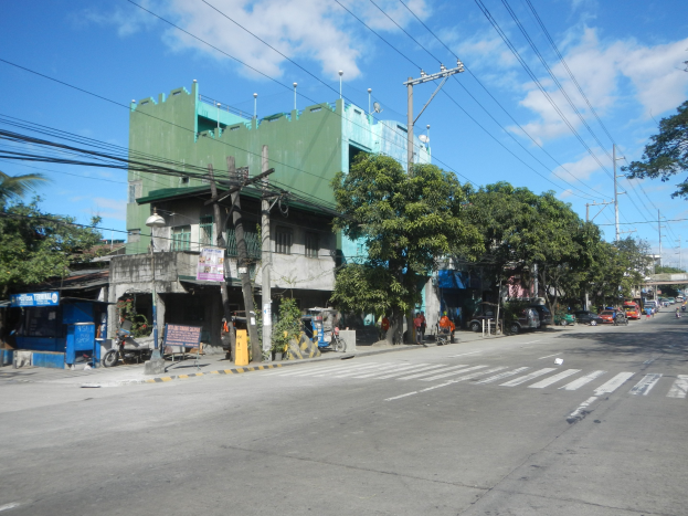 Eine Stadtstraße mit einem grünen Gebäude, fahrenden Fahrzeugen, Fußgängern auf dem Gehweg, Stromleitungen und einer bewölkten Himmel