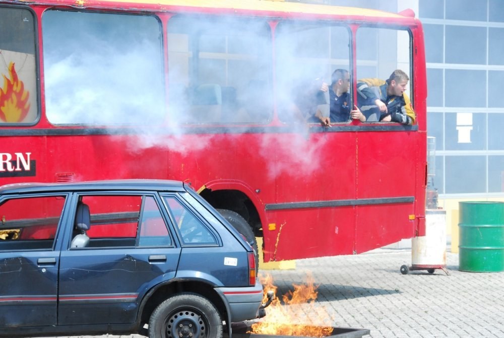 Roter Doppeldeckerbus mit Rauch, drei sichtbaren Passagieren, neben einem Auto geparkt, vor einem Gebäude mit Glasfenstern und einem Fass auf der rechten Seite.