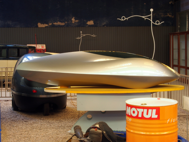 Ein kleines Hydrofoil-Boot mit schlankem, aerodynamischem Design steht auf einem Tisch neben einem Auto, mit zwei Trommeln auf dem Boden und einem Metallzaun im Hintergrund; eine Glastür und eine Tafel mit Text sind auf der rechten Seite zu sehen.