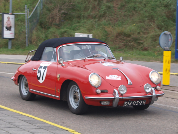 Roter Porsche 356 Speedster mit zwei Insassen auf einer Rennstrecke, neben einer am Pfosten angebrachten Tafel, mit Vegetation und einem Zaun im Hintergrund.