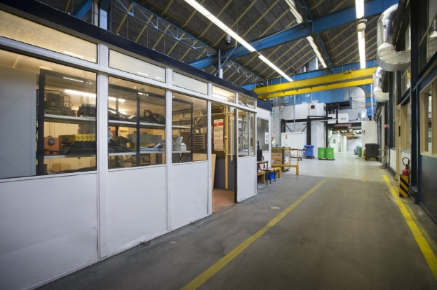 Großes Lagerinneres mit zahlreichen Fenstern, Türen, Tischen, Rohren, Lampen und anderen industriellen Objekten.