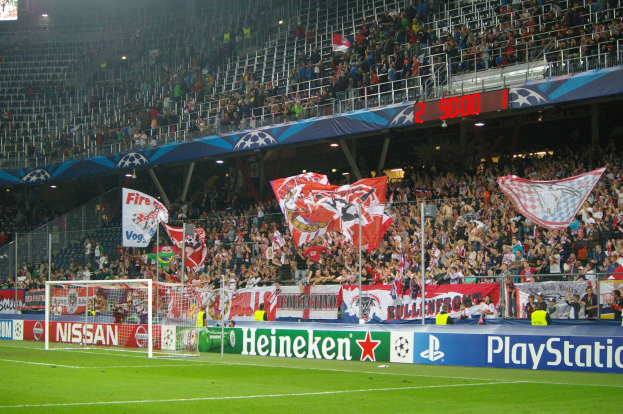 Ein Fußballspiel in einem hell erleuchteten Stadion mit einer großen Zuschauermenge, grünem Rasen, Torstangen, Bannern und einem Bildschirm in der linken oberen Ecke.