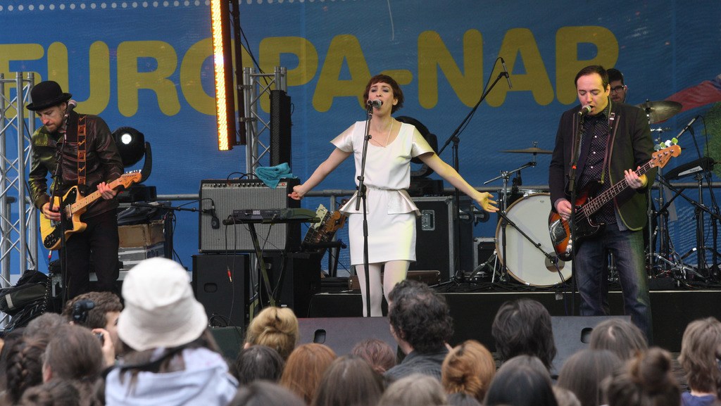 Vier Personen auf der Bühne: eine Frau in einem weißen Kleid, die singt, zwei Männer, die Gitarren spielen, und eine Menge unten mit Lautsprechern und einer Wand im Hintergrund.