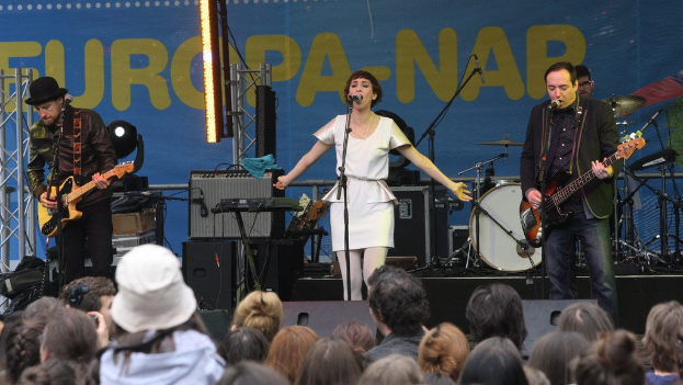 Vier Personen auf der Bühne: eine Frau in einem weißen Kleid, die singt, zwei Männer, die Gitarren spielen, und eine Menge unten mit Lautsprechern und einer Wand im Hintergrund.