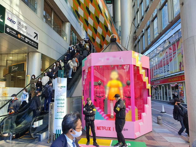 Eine Gruppe von Menschen steht vor einem Glaswand-Gebäude mit Texttafeln, einige tragen Masken, während eine Rolltreppe mit Fahrgästen zu sehen ist; eine pinkfarbene Box in der Mitte zeigt Schaufensterpuppen und trägt den Text "Netflix's new Netflix booth in Tokyo."