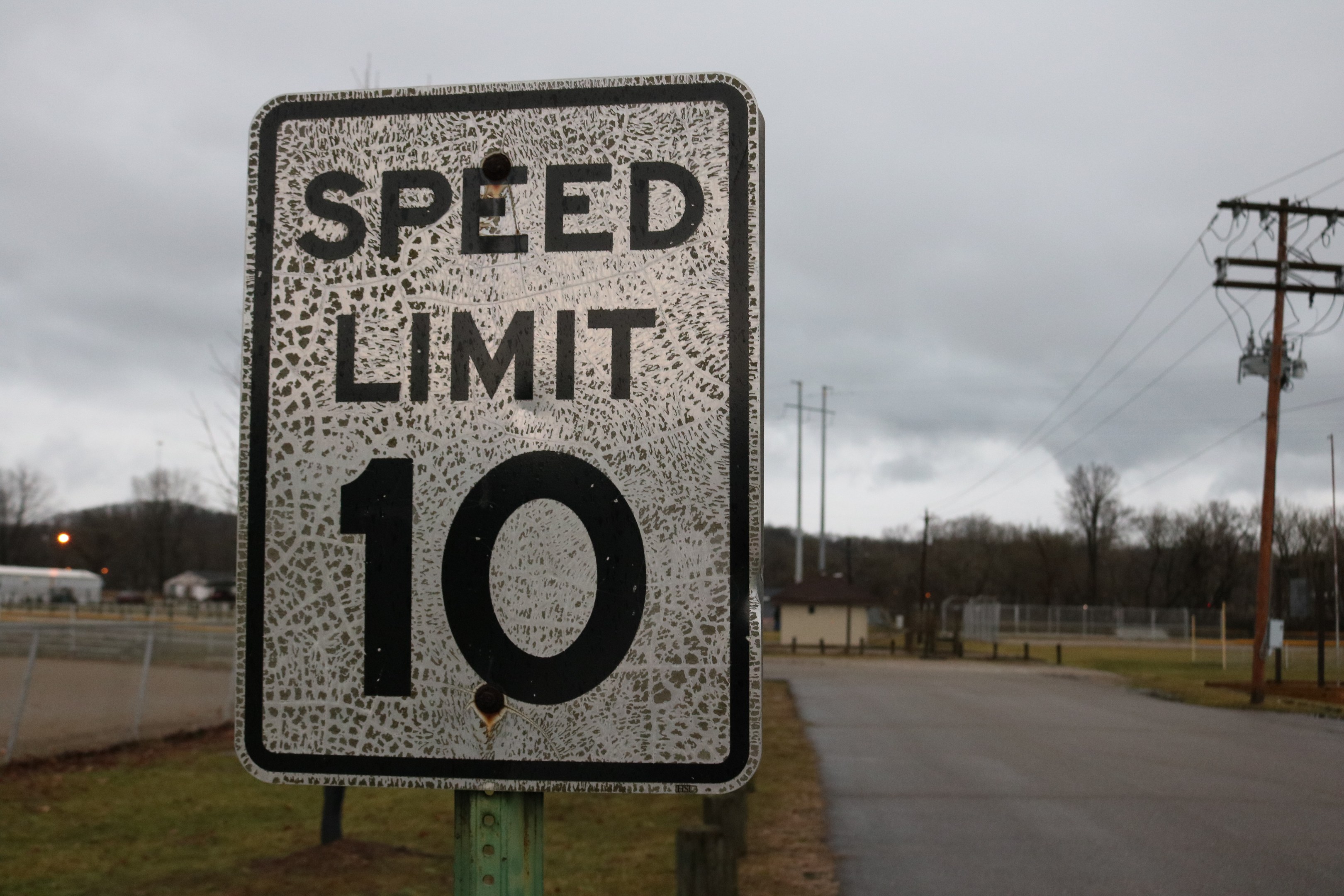 Geschwindigkeitsbegrenzungsschild am Straßenrand mit umliegenden Gras, Strommasten, Drähten, einem Zaun, Häusern, Bäumen und einem bewölkten Himmel im Hintergrund.