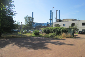 Gro├če Fabrik mit zahlreichen geparkten Autos, umgeben von Geb├Ąuden, Stra├čeninfrastruktur, Grünfl├Ąchen und einem bew├Âlktem Himmel.