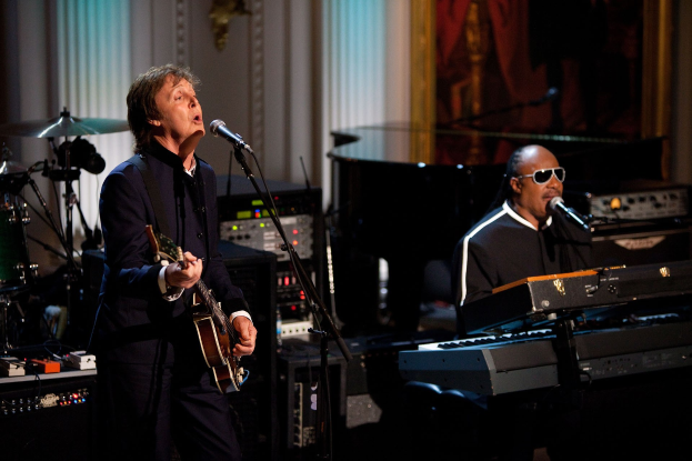 Paul McCartney spielt eine Gitarre und singt in ein Mikrofon, während Stevie Wonder, der eine Brille trägt, ein Piano auf der Bühne spielt.