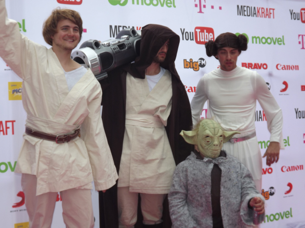 Eine Gruppe von Menschen in Star-Wars-Kostümen, die auf einem roten Teppich posen, wobei einer ein Objekt und ein Banner mit Text und Logos im Hintergrund hält.