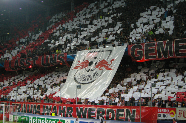 Ein volles Fußballstadion mit Sitzplätzen und Stehplätzen, Banner, Torpfosten und Flutlicht.