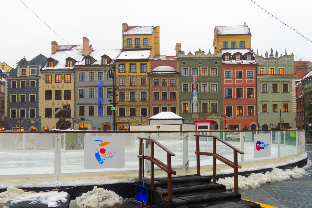 Eislaufbahn in einem Stadtplatz umgeben von Geb├Ąuden, Treppen, Gel├Ąndern, Informationsschildern, schneebedecktem Boden, Laternenmasten, B├Ąumen und einem Himmelhintergrund.