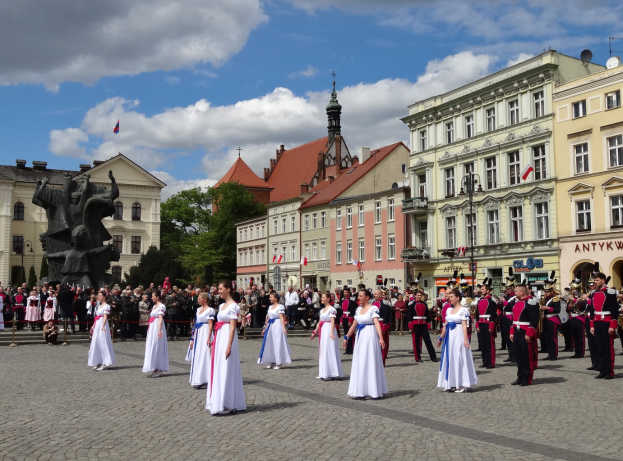 Eine Gruppe von Menschen in weißen Kleidern steht vor einer Menge mit Musikinstrumenten, mit Gebäuden, Bäumen, einer Statue, Fahnen und einem bewölkten Himmel im Hintergrund, wahrscheinlich während des Nationalfeiertagsparades der Tschechischen Republik.