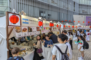Eine Gruppe von Menschen versammelt sich um einen Tisch mit Essen auf einem Food-Festival in Japan, wobei einige Gläser halten und andere das Essen untersuchen, während Schilder mit Text und Bildern, Lichter und Laternen von der Decke hängen und ein Gebäude im Hintergrund zu sehen ist.