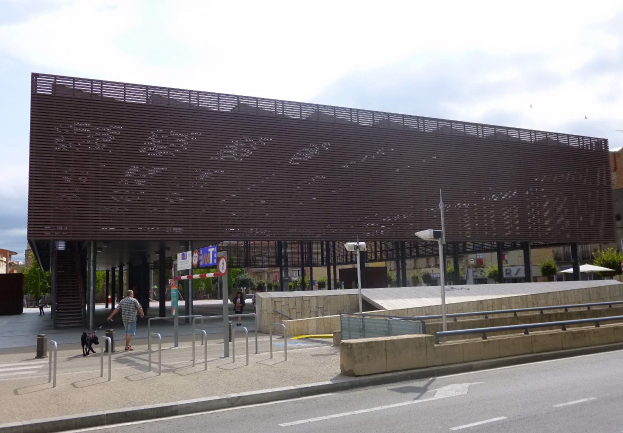 Großes modernes Gebäude mit zahlreichen Fenstern, das als neuer Eingang zur Stadt dient, umgeben von Straßeninfrastruktur, Fahrzeugen, Fußgängern, Bäumen und bewölktem Himmel.