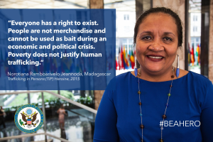 Eine Frau in einem blauen Kleid steht lachend vor einem Gebäude mit einer Aussage über Menschenhandel, mit Menschen, Pflanzen und Flaggen im Hintergrund.