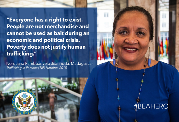 Eine Frau in einem blauen Kleid steht lachend vor einem Gebäude mit einer Aussage über Menschenhandel, mit Menschen, Pflanzen und Flaggen im Hintergrund.