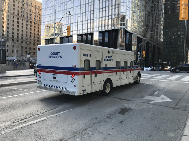 Ein Rettungswagen steht an einer Stadtstraße geparkt, umgeben von hohen Gebäuden, Straßenlaternen, Verkehrspfosten, Ampeln und Menschen auf dem Gehweg, mit dem Himmel im Hintergrund.