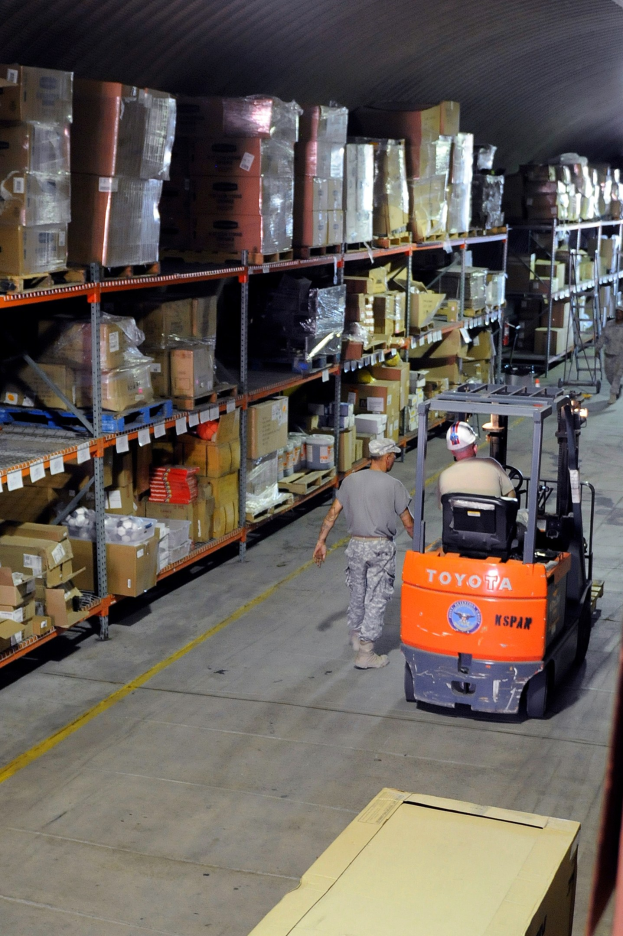 Ein Mann bedient einen Gabelstapler in einem Lagerhaus, das mit Kartonagen und anderen Gegenständen auf Regalen gefüllt ist, mit einer Kiste am unteren Bildrand.