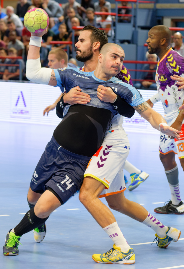 Eine Gruppe von Männern, die in einem Stadion Handball spielen, wobei einer den Ball hält, und Zuschauer im Hintergrund sitzen.