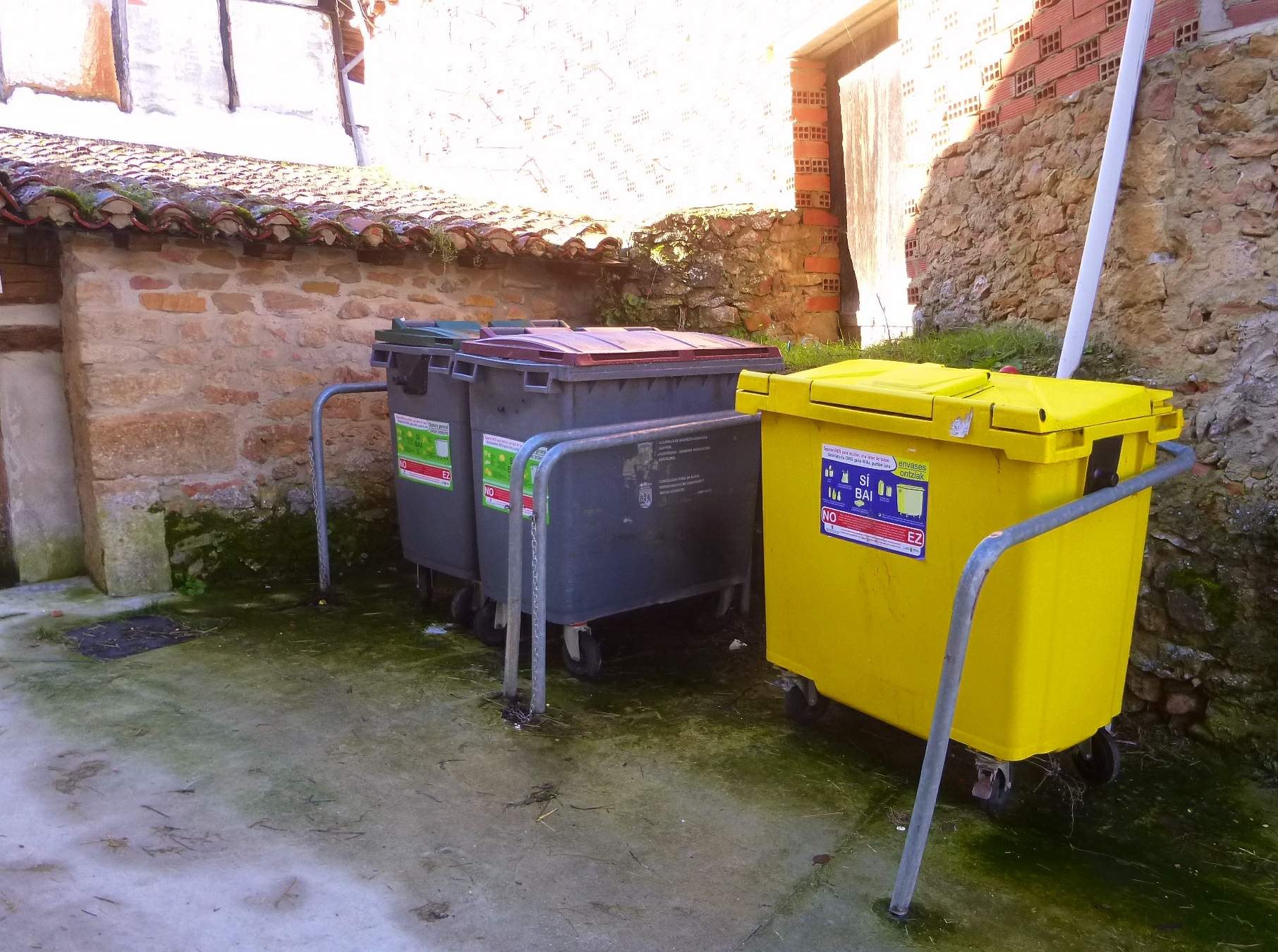Zwei gelbe und graue Recyclingcontainer mit Rädern nebeneinander vor einem Gebäude mit einer Wand und einem Fenster.