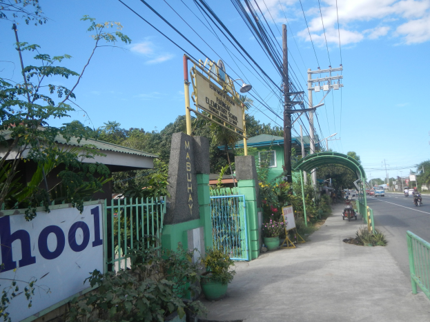 Schuleingangsschild am Straßenrand mit einem Bogen, Tor und Namensschild, umgeben von Fahrzeugen, Pflanzen, Bäumen, Strommasten mit Drähten und einem bewölkten Himmel.