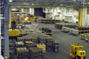 Großes Lagerinnere mit mehreren Lastwagen, Containern, Menschen, Leuchten, Metallstangen und einem sichtbaren Rad.