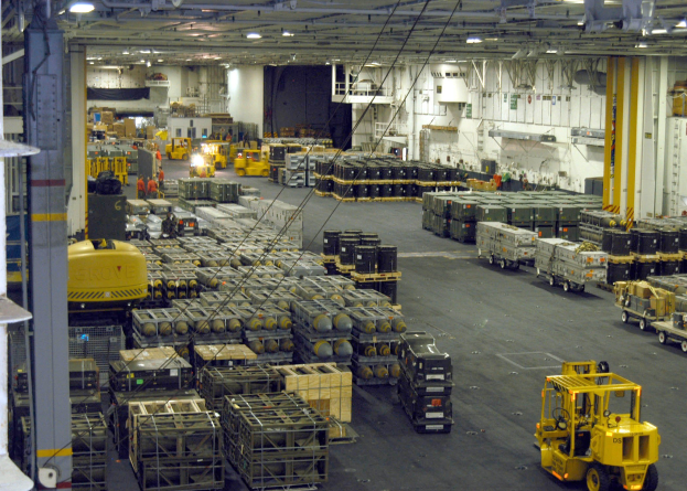 Großes Lagerinnere mit mehreren Lastwagen, Containern, Menschen, Leuchten, Metallstangen und einem sichtbaren Rad.