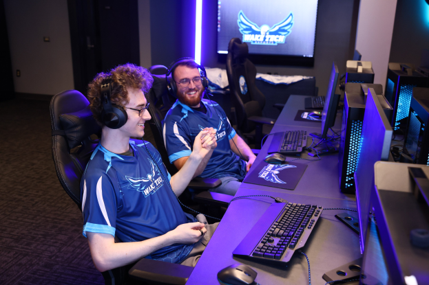 Zwei Männer in blauen T-Shirts und Kopfhörern sitzen an einem Schreibtisch mit Monitoren, Tastaturen, Mäusen und Kabeln und spielen ein Spiel vor einer wandmontierten Bildschirmwand mit dem Wack Tech-Logo und Lichtern.