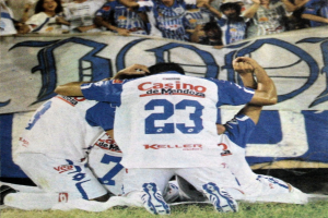 Gruppe von Fußballspielern feiert auf dem Boden vor einer jubelnden Menge mit einem Banner im Hintergrund.