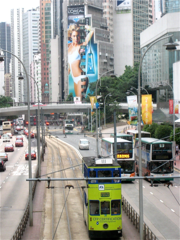 Eine belebte Stadtstraße mit verschiedenen Fahrzeugen, darunter Doppeldeckerbusse, Autos und andere Fahrzeuge, unter einem klaren blauen Himmel, umgeben von hohen Gebäuden, Pfählen, Lampen, Schildern, Bannern, Bäumen und einer Brücke, mit Text unten.