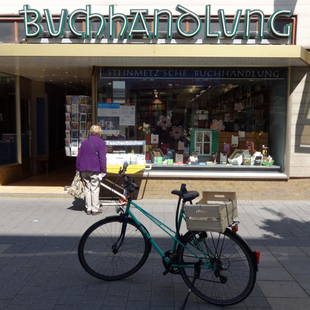 Eine Frau steht neben einem geparkten Fahrrad vor einem Laden, mit einem Fahrradrad in Vordergrund und durch die Gläser des Ladens sichtbaren Büchern.